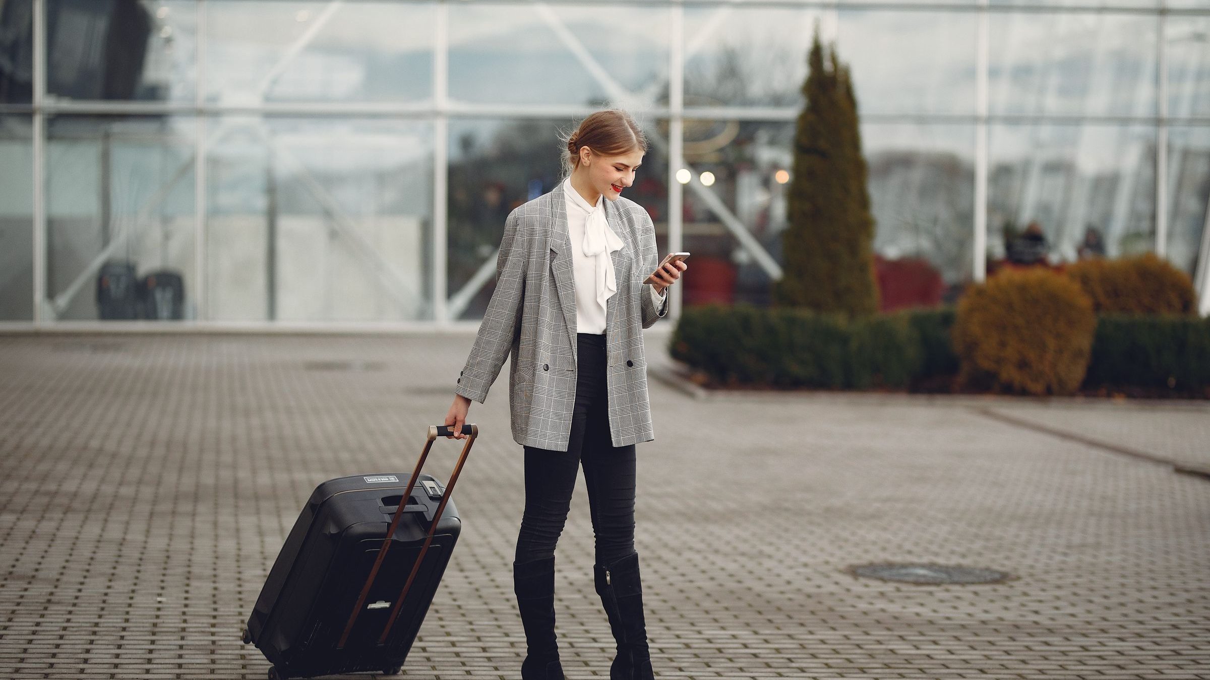 Viajante puxando a mala na frente de um terminal de aeroporto, sorrindo enquanto olha o celular