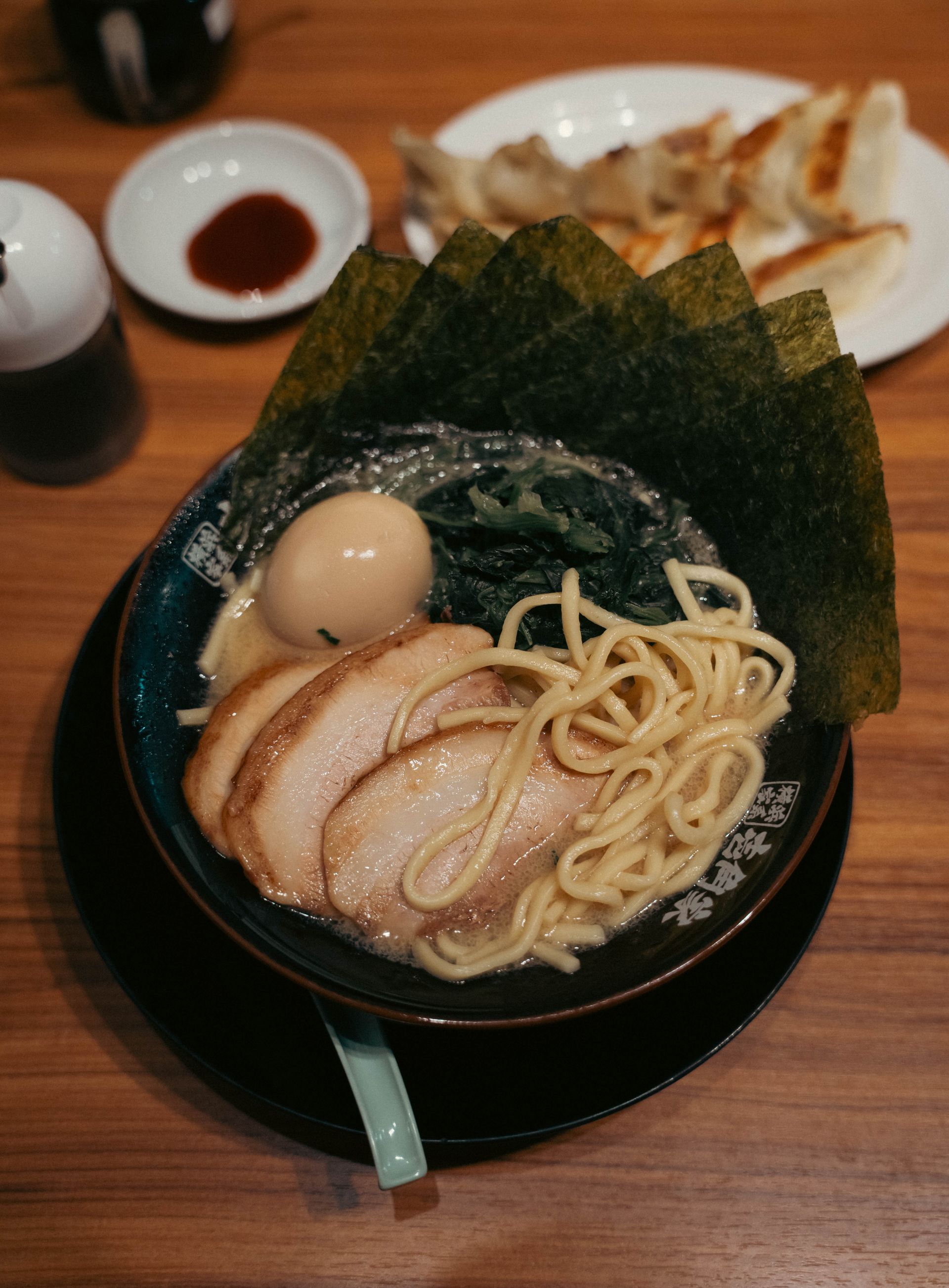 Uma tigela de ramen com chashu, ovo marinado, espinafre e folhas de nori, com gyoza ao fundo