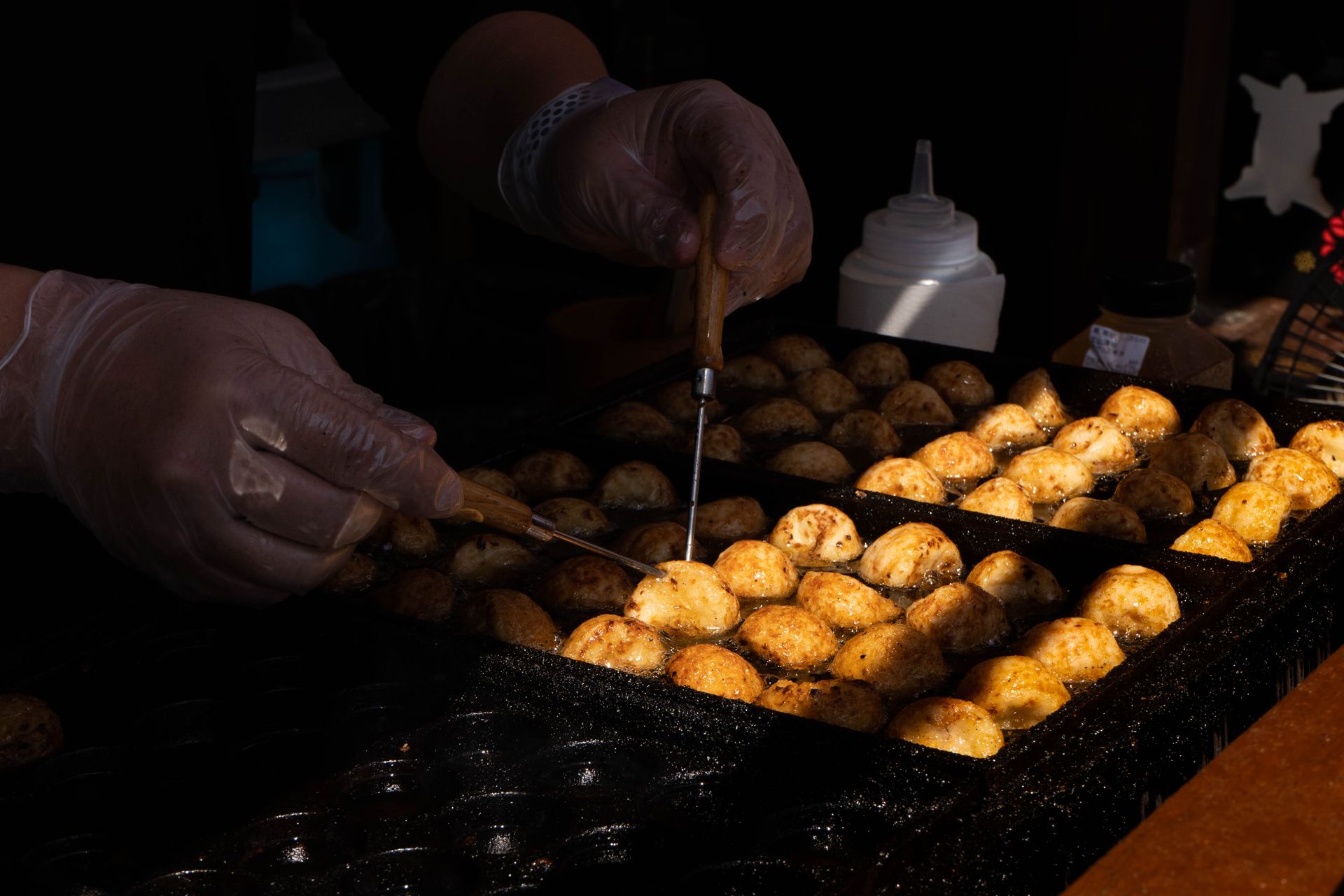 Mãos de luva viram bolinhos de takoyaki numa chapa de ferro de uma barraca de rua