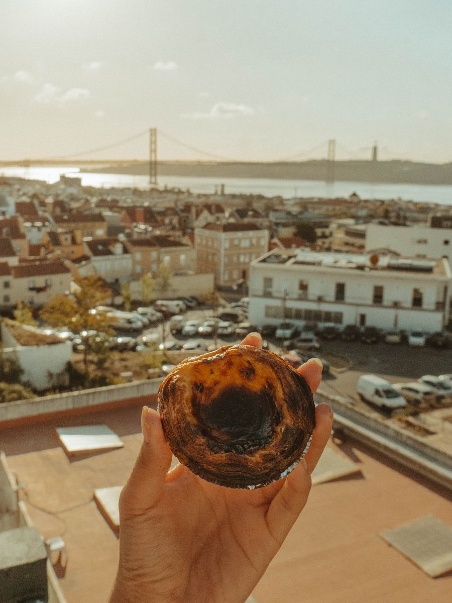 Mão segurando um pastel de nata, com Lisboa e a Ponte 25 de Abril ao fundo