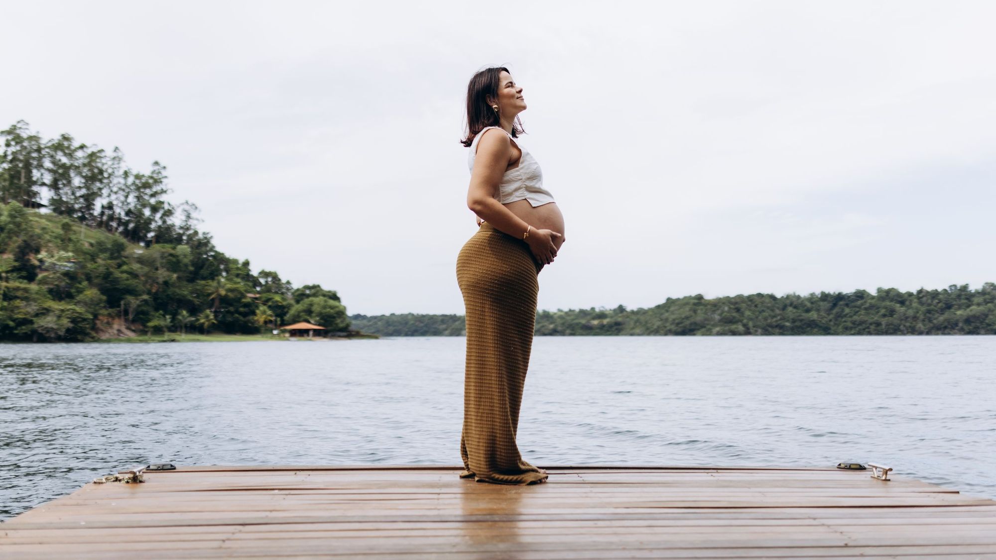Gestante de perfil num píer de madeira à beira de um lago, com as mãos na barriga